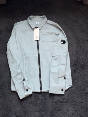 Ljusgrå overshirt från C.P. Company - Stilren ljusgrå overshirt från C.P. Company med rak passform, dragkedja framtill och klassisk krage. Bröstficka med lock och ikonisk logga på vänster ärm. Tillverkad i lätt bomullsmaterial, perfekt för lager-på-lager-stil.