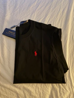 Svart t-shirt från Polo Ralph Lauren - Svart t-shirt från Polo Ralph Lauren med klassisk rund hals och det ikoniska röda broderade logot på bröstet. Tillverkad i mjuk bomull för en skön känsla och enkel stil. Perfekt till jeans eller shorts för en clean look. Pris kan diskuteras 