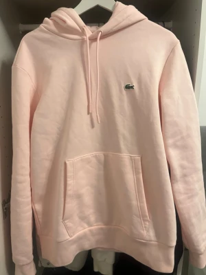 Ljusrosa hoodie från Lacoste - Säljer en fantastiskt fin ljusrosa hoodie från Lacoste. Tröjan är endast använd vid ett enstaka tillfälle då den tyvärr var lite för stor för mig. Den är därför i absolut nyskick! Hög kvalitet, klassisk passform och den ikoniska logotypen på bröstet. Ett perfekt tillfälle att fynda en nästintill oanvänd märkeshoodie.                 NY PRIS 1695kr, PRIS KAN DISKUTERAS! 