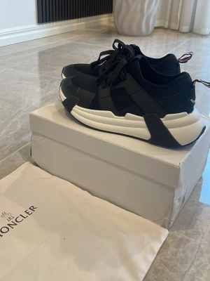 Svarta Moncler sneakers med vit sula - Snygga svarta sneakers från Moncler med chunky vit sula och coola detaljer i gummi. Skorna har Monclers klassiska logga på hälen och en röd, vit och blå flik bak. Perfekta för dig som gillar streetstyle och exklusiva märken.