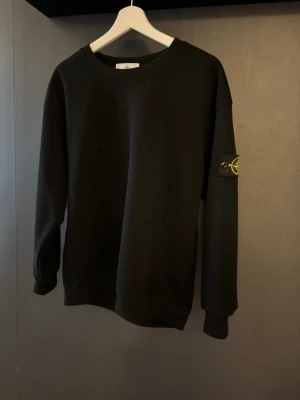  Stone Island tröja  - Säljer en svart sweatshirt från Stone Island med den ikoniska loggan på vänster ärm. Tröjan har rund halsringning, långa ärmar och ribbade muddar. Perfekt för en clean och stilren look.
