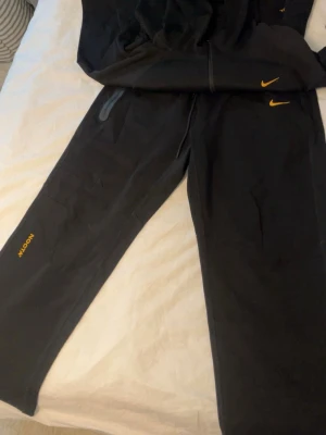 Nike svart loungewear-set NOCTA - Svart loungewear-set från Nike NOCTA med gul logga på både hoodie och byxa. Hoodien har dragkedja, huva och fickor med dragkedja. Byxorna har resår i midjan och NOCTA-tryck i gult på benet. Perfekt för chill eller streetwear.
