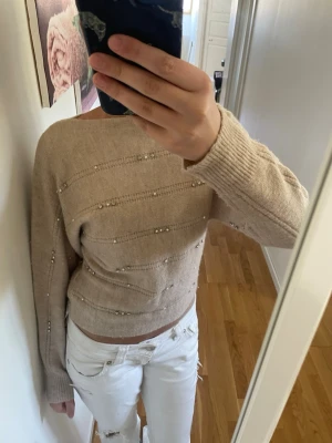 Beige stickad tröja med pärldetaljer - Trendig beige stickad tröja med pärldetaljer och diskreta ränder. Tröjan har en avslappnad passform, långa ärmar och bred ribbad mudd vid ärmslut och nederkant. Perfekt för dig som gillar en stilren men ändå unik look.