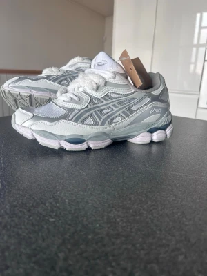 ASICS vita och grå sneakers - Snygga sneakers från ASICS i vitt och ljusgrått med silvriga detaljer. Skorna har en sportig design med mesh och syntetmaterial, samt en chunky sula för extra komfort. Perfekta för dig som gillar en clean och modern stil.