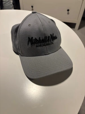 Grå keps från Mitchell & Ness - Snygg grå keps från Mitchell & Ness med svart broderad logga framtill och justerbar rem bak. Klassisk böjd skärm och stilren design, perfekt för dig som gillar streetwear och sportig stil. Tillverkad i slitstarkt tyg med detaljer på insidan.