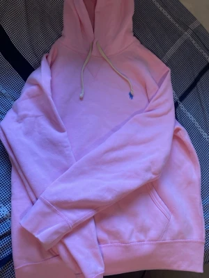 Rosa hoodie från Polo Ralph Lauren - Snygg ljusrosa hoodie från Polo Ralph Lauren med klassisk huva och vita dragsnören. Broderad blå logga på bröstet och känguruficka framtill. Mjuk bomullsblandning som är skön att ha på sig. Perfekt för en avslappnad och trendig look. Storlek M helt oanvändt, köptes från Zalando för en månad sen finns kvitto!
