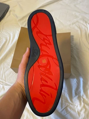 Mörkblå Louboutin sneakers i mocka - Snygga mörkblå sneakers från Christian Louboutin med klassisk röd sula och ovandel i mjuk mocka. Skorna har rund tå, svarta skosnören och diskret branding på sulan. Perfekta för dig som vill sticka ut med exklusiva detaljer och lyxig känsla.