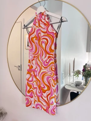 Mönstrad halterneckklänning i orange och rosa - Säljer en kort halterneckklänning med färgstarkt psykedeliskt mönster i orange, rosa och vitt. Klänningen har en åtsittande passform och är ärmlös, perfekt för sommarens alla tillfällen. Materialet känns lätt och svalt.