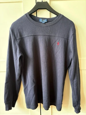 Mörkblå långärmad tröja Polo Ralph Lauren barn (14-16 år) - Mörkgrå långärmad tröja från Polo Ralph Lauren med röd broderad logga på bröstet. Tröjan har rund halsringning och är tillverkad i ett mjukt, strukturerat material som känns skönt mot huden. 