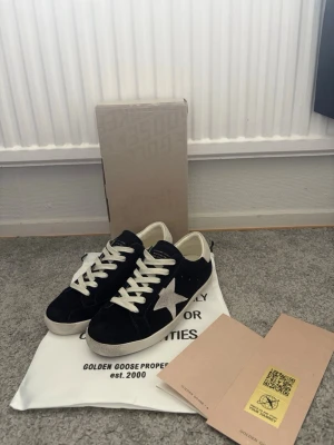 Golden Goose svarta sneakers med stjärna - Svarta sneakers från Golden Goose med vit stjärndetalj på sidan, vita skosnören och vit sula. Skorna har en klassisk låg modell och är tillverkade i mocka och läder. Perfekta för dig som gillar en stilren men ändå unik look.