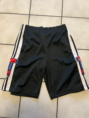 Svarta Nike shorts med knappar storlek S/M - Säljer ett par svarta Nike shorts med vita och blå ränder längs sidorna samt tryckta Nike-loggor i rött och blått. Shortsen har tryckknappar längs benen och elastisk midja för skön passform. Perfekta för en sportig look.