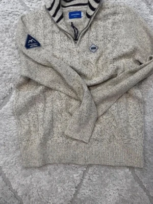 Grå stickad tröja från Jack & Jones - Snygg gråmelerad stickad tröja från Jack & Jones med halv dragkedja och ståkrage med marinblå ränder. Tröjan har patchdetaljer på bröstet och ärmen, samt ribbstickade muddar. Perfekt för kyliga dagar.