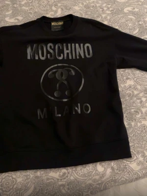 Svart sweatshirt från Moschino - Cool svart sweatshirt från Moschino med glansigt tryck framtill och texten 'Milano'. Tröjan har rund halsringning, långa ärmar och ribbade muddar. Perfekt för en trendig och avslappnad stil.