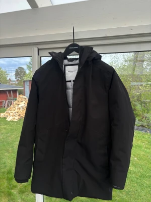 Svart parkas från Jack & Jones - Stilren svart parkas från Jack & Jones med huva och dragkedja samt tryckknappar framtill. Jackan har en lätt vadderad insida och raka ärmar. Perfekt för kyliga dagar och enkel att matcha med olika outfits.