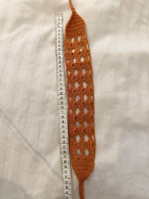 Handvirkad orange hårband - Unikt handvirkat hårband i en varm orange färg. Hårbandet har ett luftigt, hålmönstrat design och är ca 37 cm långt och 6 cm brett. Perfekt accessoar för att ge din frisyr en bohemisk touch. Tillverkat i mjukt bomullsgarn.