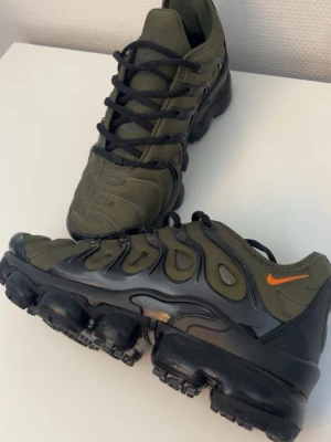 Nike Air Vapormax Plus olivgrön - Nike Air Vapormax Plus sneakers i olivgrönt tyg med svarta detaljer och en orange swoosh på sidan. Skorna har en chunky svart sula med synliga luftbubblor och svarta snören. Perfekt för dig som gillar streetwear och sportig stil.