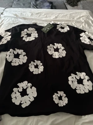 Svart t-shirt med blommigt tryck Denim Tears - Svart t-shirt från Denim Tears med stora vita blommiga tryck över hela tröjan. Klassisk passform och rund halsringning. Perfekt för dig som vill sticka ut med en unik och trendig look.