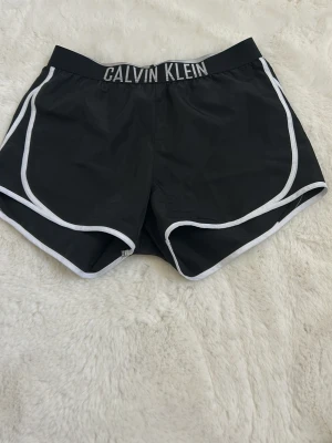 Svarta Calvin Klein shorts med vit kant - Snygga svarta shorts från Calvin Klein med vit kant och bred resår i midjan där loggan syns tydligt. Perfekta för sommarens alla aktiviteter och tillverkade i ett lätt syntetmaterial som känns bekvämt och sportigt.