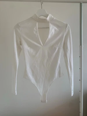 Vit långärmad body med polokrage - Stilren vit body med lång ärm och polokrage. Djup v-ringning framtill ger en trendig look. Tillverkad i stretchigt material för en bekväm passform. Perfekt att styla med jeans eller kjol för en snygg outfit.