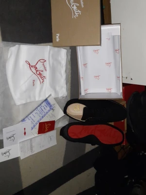 Svarta Louboutin sneakers med röd sula - Snygga svarta sneakers från Christian Louboutin med den ikoniska röda sulan och diskret logga på sidan. Skorna har en klassisk låg siluett och är tillverkade i mocka med svarta skosnören. Perfekta för dig som vill sticka ut med exklusiv stil.