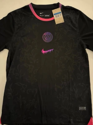 Svart PSG x Nike t-shirt med mönster - Cool svart t-shirt från Nike i samarbete med PSG. Tröjan har ett diskret blommönster, lila och rosa detaljer vid kragen samt PSG-logga och Nike swoosh framtill. Tillverkad i lätt och ventilerande material, perfekt för träning eller streetwear.