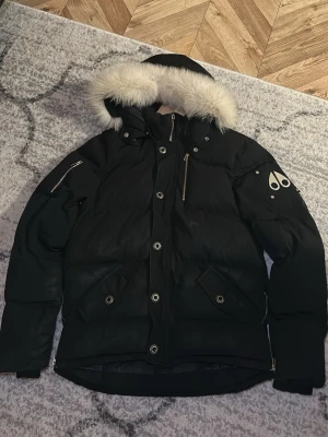 Svart dunjacka från Moose Knuckles - Snygg svart dunjacka från Moose Knuckles med avtagbar huva och vit fuskpäls. Jackan har flera fickor med dragkedja och tryckknappar samt logga på ärmen. Perfekt för kalla dagar och riktigt varm.