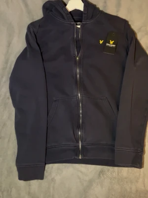 Mörkblå hoodie från Lyle & Scott - Mörkblå hoodie med dragkedja från Lyle & Scott Junior. Klassisk design med huva, känguruficka och gul broderad logga på bröstet. Mjuk bomullskänsla och stilren look som passar perfekt till jeans eller joggers.