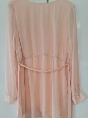 Ljusrosa transparent klänning med knytband - Söt ljusrosa klänning i transparent chiffong med långa ärmar och knappar vid ärmsluten. Blusen har ett  knytband i midjan. Innerklänning