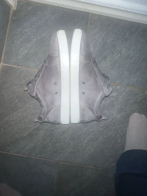 Christian Louboutin grå mocka sneakers - Säljer ett par gråa sneakers från Christian Louboutin med klassisk röd sula och snörning. Skorna är i mocka med vit platt sula och rund tå. Medföljer originalkartong, dustbag och äkthetskort. Perfekt för dig som gillar exklusiva märken och snygg design. I använd skick