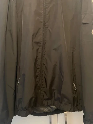 Svart vindjacka med luva - Snygg svart vindjacka med dragkedja framtill, två sidofickor med dragkedja och en ficka på ärmen. Jackan har en praktisk huva och elastisk nederkant. Tillverkad i lätt och tåligt material, perfekt för blåsiga dagar.