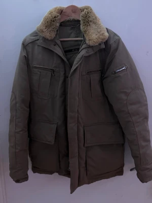  Hetregó jacka med päls - Snygg beige parkas från Hetregó med mjuk fuskpäls runt kragen. Jackan har flera praktiska fickor med dragkedja och lock, quiltat innerfoder och en klassisk rak passform. Perfekt för kalla dagar när du vill hålla stilen.