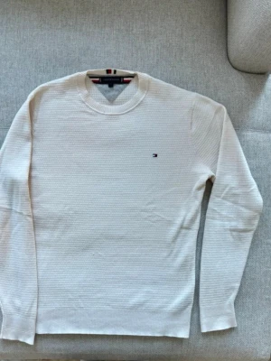 Vit stickad tröja Tommy Hilfiger - Vit stickad tröja från Tommy Hilfiger med diskret logga broderad på bröstet. Tröjan har rund halsringning, lång ärm och en strukturerad stickning som ger en stilren look. Perfekt för lager-på-lager och enkel att matcha.