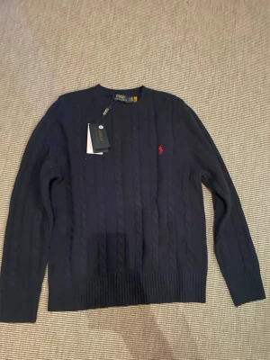 Mörkblå kabelstickad tröja Polo Ralph Lauren - Mörkblå kabelstickad tröja från Polo Ralph Lauren med rund halsringning och röd broderad logga på bröstet. Tröjan har långa ärmar och ribbade muddar vid ärmslut och nederkant. Perfekt för en stilren och klassisk look.