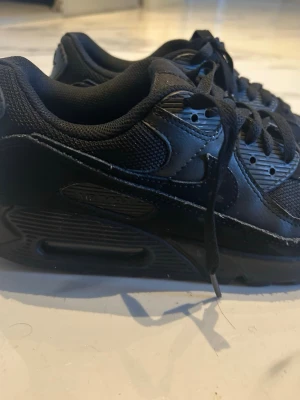 Svarta Nike Air Max 90 sneakers - Säljer ett par helsvarta Nike Air Max 90 sneakers med klassisk chunky sula och synlig Air-enhet. Skorna har ovandel i både mesh och läder, samt svarta skosnören och diskreta detaljer. Perfekta för dig som gillar en clean och sportig look. Jag har endast haft på mig dem ett antal gånger(4 gånger).