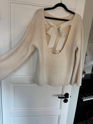 Krämvit stickad tröja med rosett - Supersöt krämvit oversize stickad tröja från Gina Tricot med djup rygg och stor dekorativ rosett i ryggen. Tröjan har ribbstickat material, långa ärmar och en mjuk, mysig känsla. Perfekt för lager-på-lager och att styla med jeans eller kjol. 