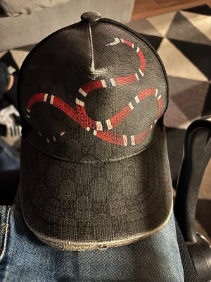 Svart Gucci-keps med ormtryck - Snygg svart keps från Gucci med ett fett ormtryck i rött, vitt och svart på framsidan. Kepsen har klassisk böjd skärm och mesh på insidan med Gucci-loggor. Materialet känns lyxigt och detaljerna är riktigt stilrena.