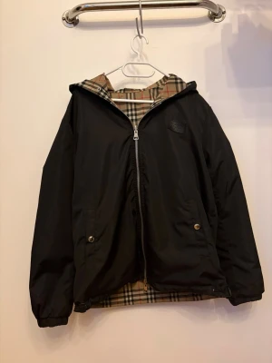 Svart Burberry vindjacka med huva - Stilren svart vindjacka från Burberry med huva och klassiskt rutigt foder. Jackan har dragkedja framtill, två fickor med tryckknappar och Burberry London England-logga på bröstet. Perfekt för att hålla vinden borta och ge en snygg look.