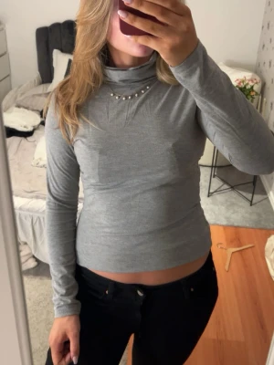 Grå polotopp med pärldetaljer - Snygg grå polotopp med långa ärmar och pärldetaljer vid halsen. Toppen har en slim passform som framhäver figuren. Perfekt att matcha med höga jeans eller kjol.