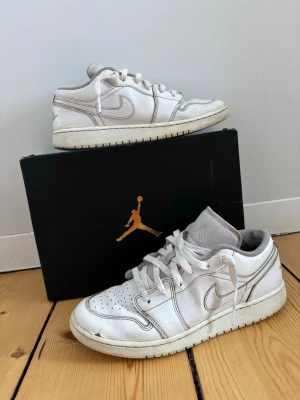 Vita Air Jordan 1 Low sneakers - Säljer ett par vita Air Jordan 1 Low sneakers med klassisk siluett och perforerad tåbox. Skorna har vita läderpaneler, vita skosnören och den ikoniska swoosh-loggan på sidan. Hör av er vid frågor och funderingar!