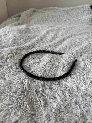 Svart diadem med glittrande stenar - Säljer ett svart diadem dekorerat med små glittrande stenar som ger en snygg och elegant look. Diademet är smalt och har en klassisk rund form. Perfekt för att lyfta din frisyr med lite extra shimmer. Använt fåtal gånger!
