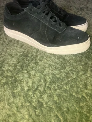 Svarta sneakers från Axel Arigato - Stilrena svarta sneakers från Axel Arigato i mocka med vit sula. Klassisk låg modell med snörning och diskret design som passar till det mesta. Perfekta för dig som gillar minimalistisk stil och vill ha ett par schyssta sneakers i garderoben.