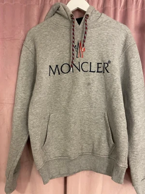 Grå Moncler hoodie med logga - Snygg grå hoodie från Moncler med stor broderad logga på bröstet och coola snören i huvan. Klassisk känguruficka framtill och ribbade muddar vid ärmar och nederkant. Perfekt för dig som vill ha en stilren och exklusiv look. Finns en fläck på tröjan (se bild)