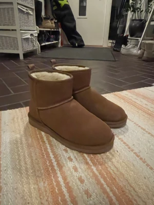 Bruna fårskinnstofflor i mocka - Mysiga bruna tofflor i klassisk UGG-stil med utsida i mocka och fluffigt foder. Modellen är ankelhög med rund tå och bred passform. Perfekta för att hålla fötterna varma ute under kalla dagar.använda två gånger 