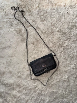 Väska  - Snygg svart axelväska från Zadig & Voltaire i strukturerat läder. Väskan har silverfärgade metalldetaljer, kedjerem och ett emblem med vingar på framsidan. Praktisk dragkedja baktill och dubbla spännen framtill.  Säljer då den inte kommer till användning, nyskick inga defekter eller skador. Nypris 2900 kr pris kan absolut diskuteras😊