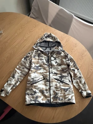 Skaljacka / Skidjacka Peak Performance  stl L (mer som M) - Nyskick! Använd 1 gång. Skaljacka och Skidjacka  vindjacka i mönster i beige, vitt och grått. Jackan har dragkedja framtill, två snedställda fickor med dragkedja och justerbar huva. Ficka framtill med duk för att putsa glasögon etc. Avtagbart snölås i midjan. Materialet är lätt och vattenavvisande.  