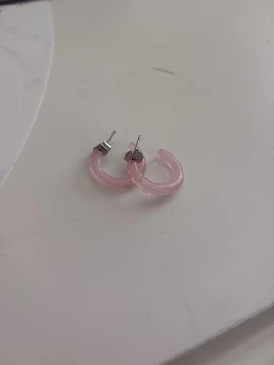 Rosa hoops örhängen i plast - Säljer ett par snygga hoops örhängen i ljusrosa plast. De har en halvcirkelformad design och är perfekta för att ge din look en söt touch. Passar dig som gillar trendiga accessoarer och vill sticka ut lite extra. Passar fint på sommarn, aldrig använt från Kappahl 