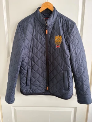 Mörkblå quiltad jacka från Morris - Quiltad mörkblå jacka från Morris med gul broderad logotyp och brittisk flagga på bröstet. Jackan har hög krage, dragkedja framtill och två sidofickor. Perfekt för dig som gillar klassisk stil och vill ha något som sticker ut lite. Nypris: 2199