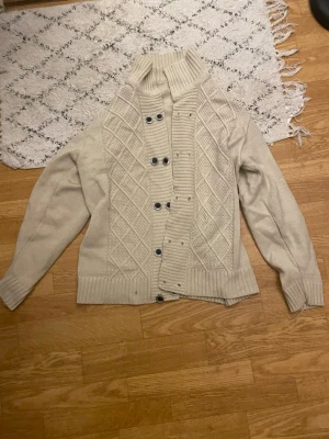 Beige stickad kofta med knappar - Snygg beige stickad kofta med hög krage och dekorativa stickade mönster framtill. Koftan har långa ärmar, ribbade muddar och knäpps med svarta knappar. Perfekt för lager på lager och en chill look.