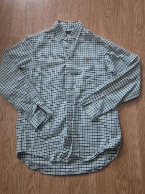 Rutig skjorta från Ralph Lauren - Klassisk rutig skjorta från Ralph Lauren i grönt, blått och vitt. Skjortan har lång ärm, knappar framtill och den ikoniska broderade loggan på bröstet. Perfekt för en stilren och avslappnad look.