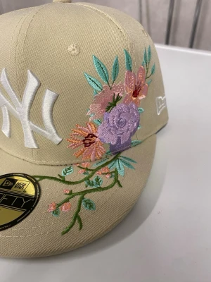 Beige New Era NY keps med blommor - Snygg beige keps från New Era med New York Yankees-logga framtill i vitt. Kepsen har broderade blommor i lila, rosa, orange och grönt på sidan och skärmen. Modellen är 59FIFTY med rak skärm och klassisk passform. Perfekt för dig som vill sticka ut med en unik accessoar.